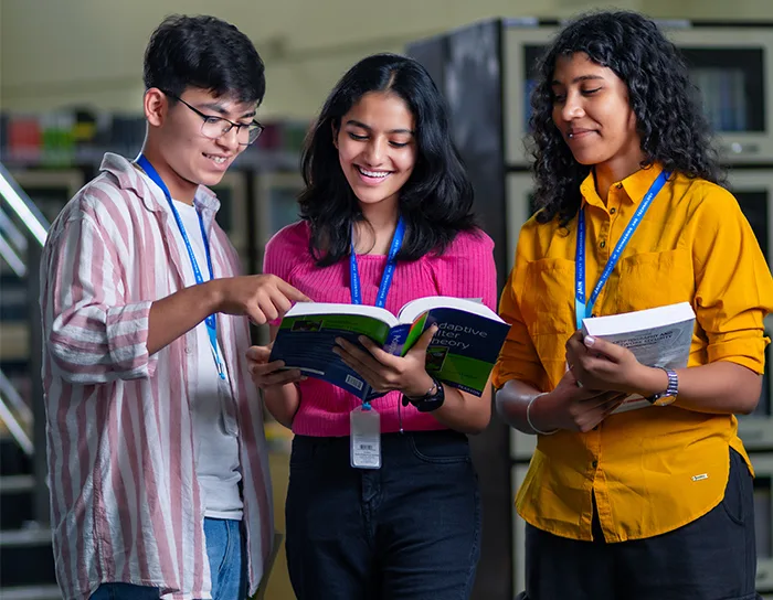Three students in a library explore a book, showcasing teamwork at BTech Aeronautical Colleges in Bangalore.