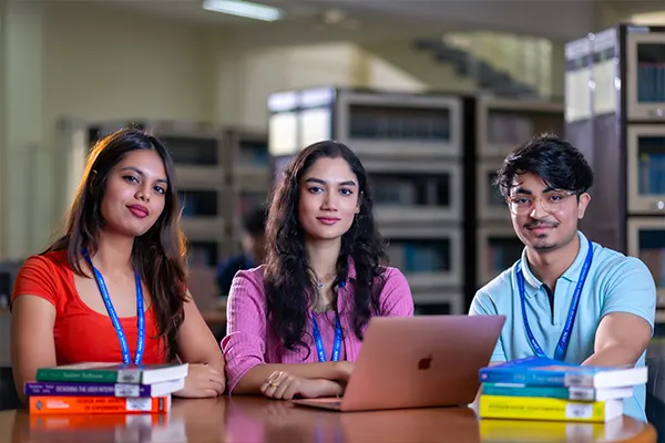 Three students smile at a table, showcasing aerospace engineering courses in India with books and a laptop.